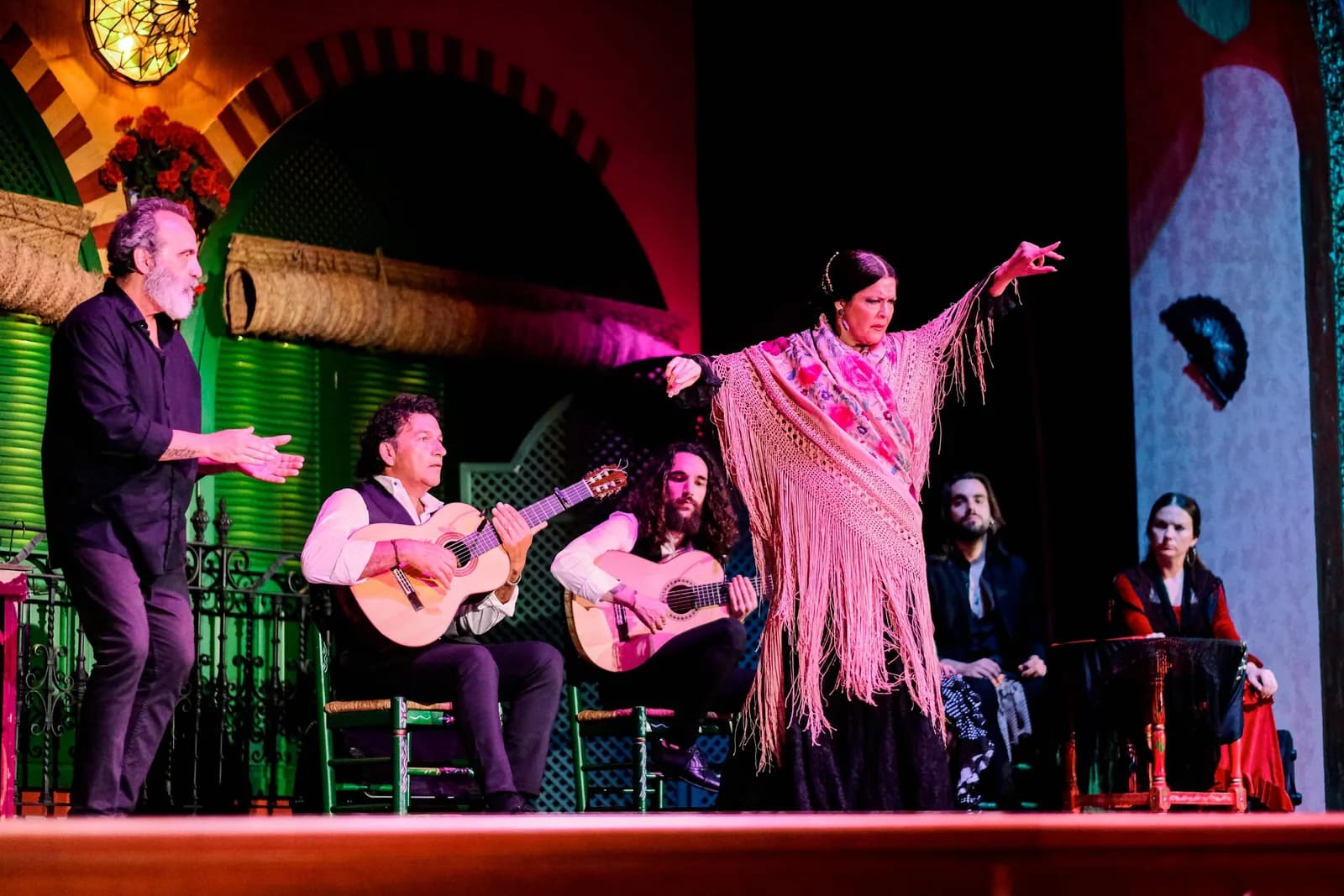 Bailaora con los brazos extendidos y palillo en escena en El Palacio Andaluz, junto a dos guitarristas y un cantaor sentados.
