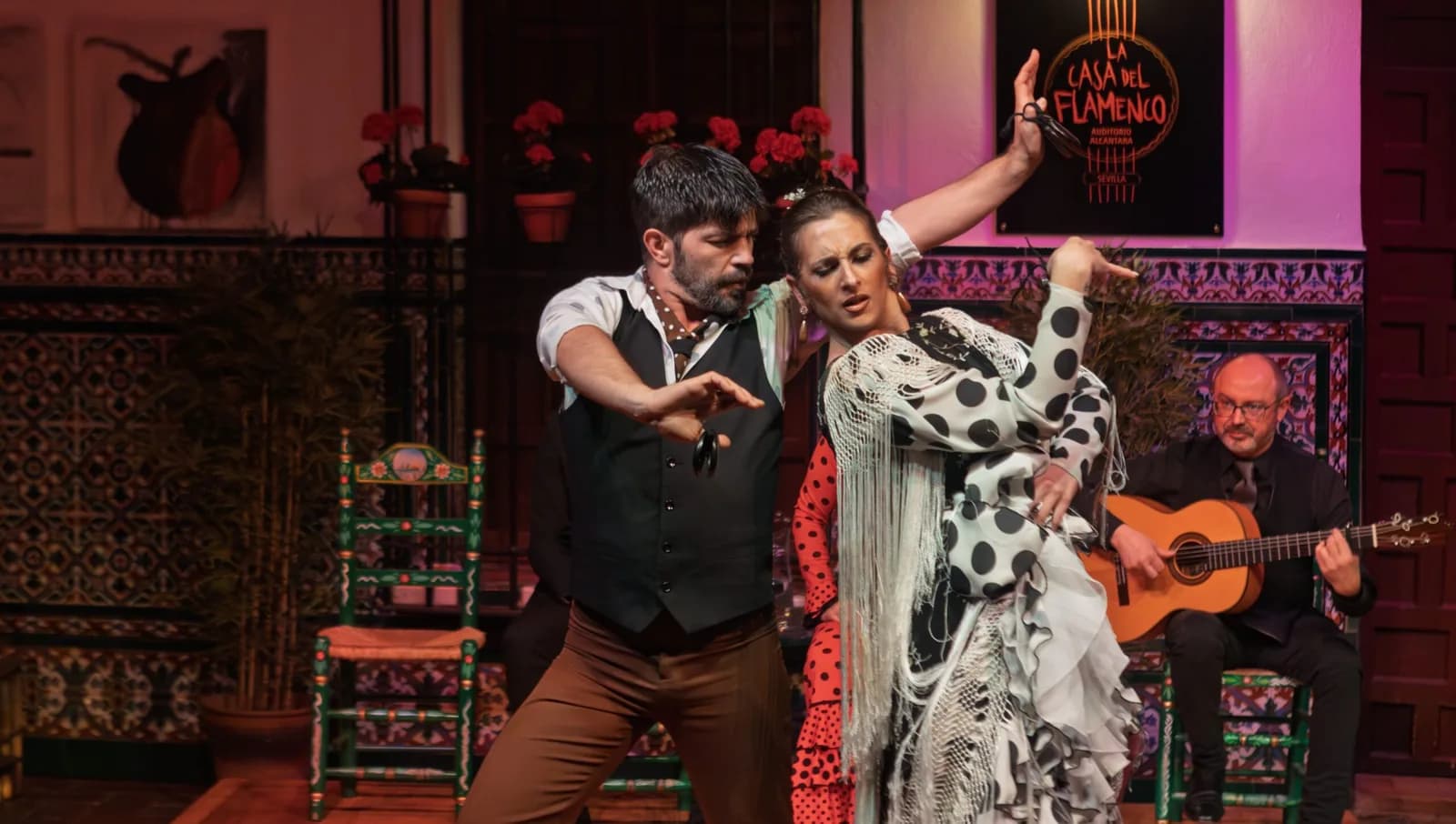 Bailaora y bailaor actuando juntos en el escenario de La Casa del Flamenco, con guitarrista al fondo bajo un letrero de neon.