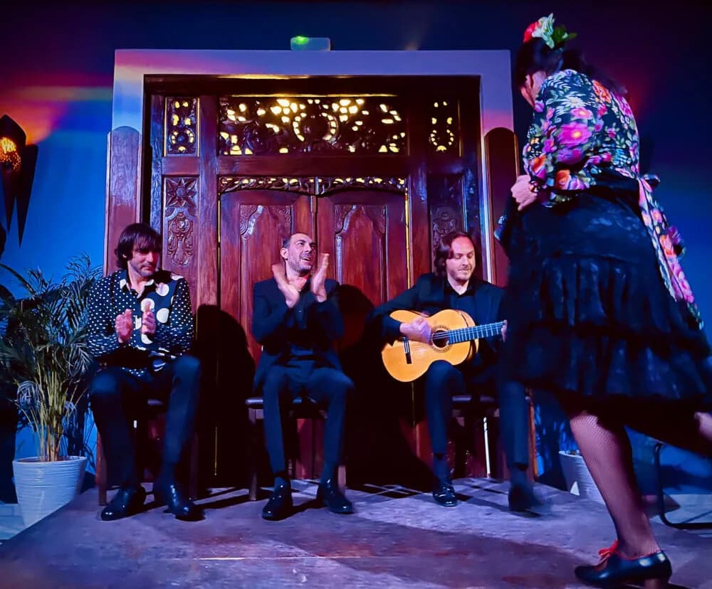 Bailaora con vestido de flores entrando al escenario de Sala Almoraima junto a tres guitarristas sentados.